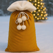A gold velvet fabric gift bag with a white faux fur topper and rope drawstring with pom-poms.  